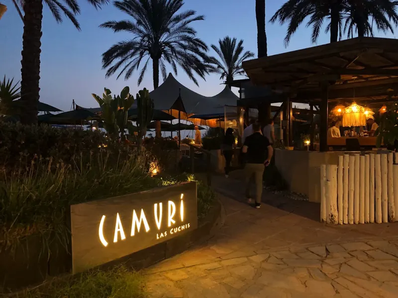 The illuminated Camurí sign at the entrance to Las Cuchis at dusk, under palm trees